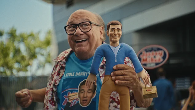 Danny DeVito is pulling the string on the back of the Eli Manning doll.